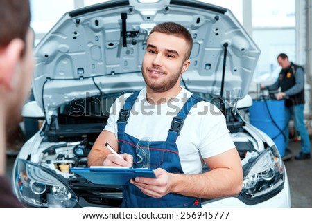 Confident mechanic. Portrait of a handsome car mechanic speaking with his client and putting information in his clipboard in the car repair station
