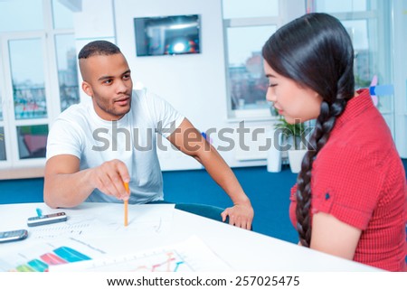 Creative team at work. Young African man and Asian woman in smart casual discussing infographics together while sitting in creative space