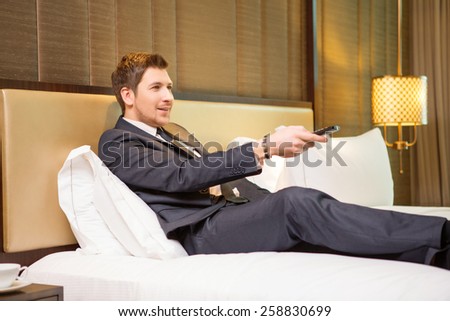 Having a break. Confident businessman in suit using remote control while sitting in the room of luxury hotel on the bed