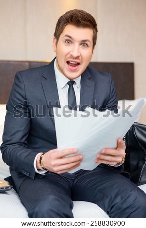 Emotional businessman. Confident young businessman in suit and tie reading documents and looking shocked while sitting on the bed in luxury hotel room