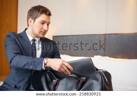 Business on the go. Confident young businessman in suit and tie putting his laptop in the suitcase while sitting on the bed in luxury hotel room