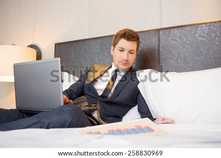 Working in hotel room. Confident young businessman in suit and tie working on laptop while sitting on the bed in hotel room and looking at analytics charts