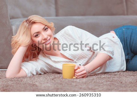 My morning coffee. Young smiling woman in casual clothing lying on the couch and holding a cup