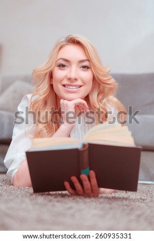 She loves reading. Top view of beautiful female student reading a book while lying on the carpet and smiling at camera