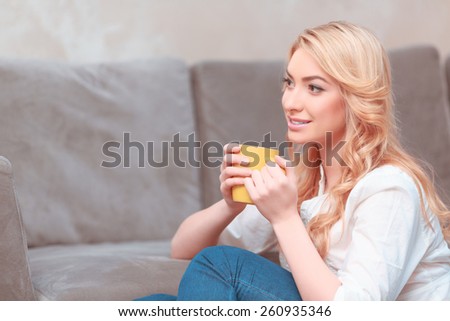 Daydreaming. Young smiling woman in casual clothing sitting by the couch and holding a cup while looking away