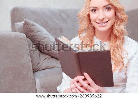 She loves reading. Beautiful female student reading a book while lying on the carpet and smiling at camera