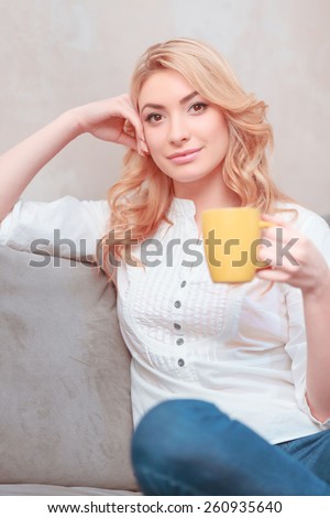 My morning coffee. Young smiling woman in casual clothing sitting on the couch and holding a cup