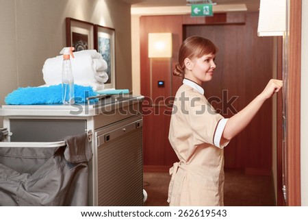 Time for room service. Smiling hotel maid in beige uniform knocking on the hotel room for room service by the maid trolley in the hotel hall