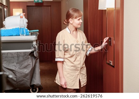 Time for room service. Smiling hotel maid in beige uniform checks climate indicators in the hall of the hotel standing just by the maid trolley