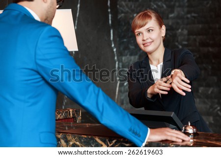 What is local time. Selective focus of an attractive hotel receptionist pointing to her watch and showing local time to the male customer in business suit leaning against the counter