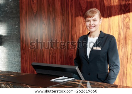Beautiful stylish hotel receptionist standing behind the service desk in a hotel lobby looking at a guest with a friendly smile