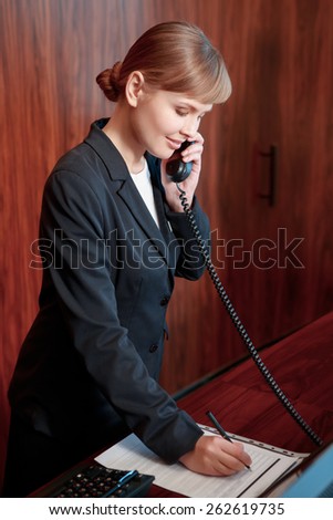 Booking confirmed. Young attractive female receptionist answering the call and making notes at the notepad by the reception desk