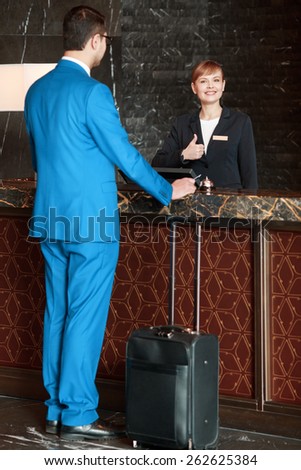 Meeting a guest. Full length selective focus view of the charming female receptionist showing her thumb up and meeting a guest businessman in blue suit with the suitcase below