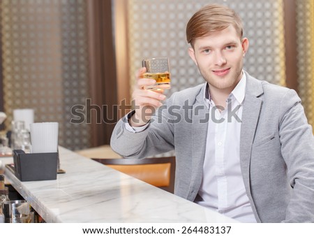 Cheers to all friends. Young smiling man toasting a glass of whiskey sitting at a counter of the bar