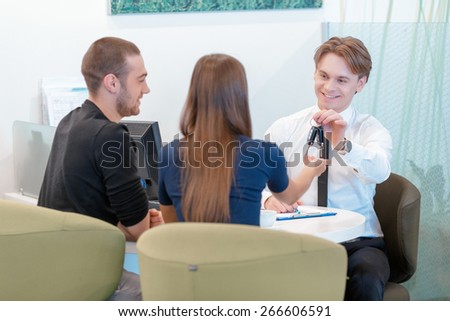 Now you are a happy owner. Front view on a smiling salesman giving keys to a happy female owner together with her couple sitting by the table backward to the camera