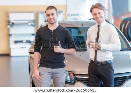 Best service ever. Male customer showing his thumb up and young smiling sales consultant pointing to the customer show delight about the purchase of a new car with a showroom view on the background