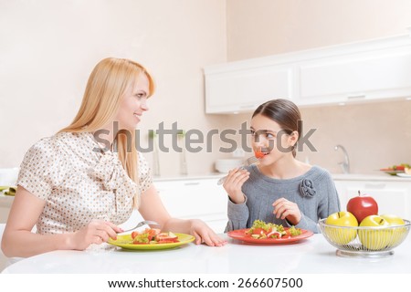 Lunchtime conversation. Charming cheerful mother and pretty daughter talking and eating salad at lunch in the kitchen
