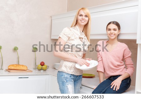 Beautiful family. Attractive blond mother and brunette daughter smiling at the camera and drying dishes at the kitchen