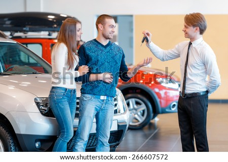 Buying their first car together. Handsome smiling sales consultant in car dealership gives keys from new car to young couple