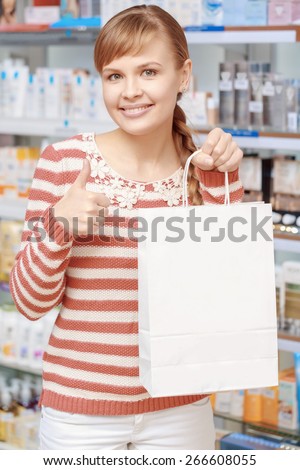 Best choice of medicines. Young cheerful woman shows thumbs up holding a paper bag copyspace in a pharmacy shop