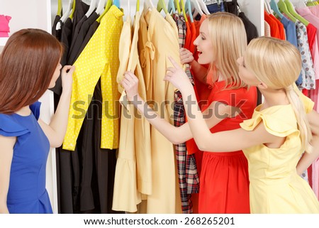Shades of color. Three young women joyfully choosing yellow shirts and dresses on a clothing rack in a shop