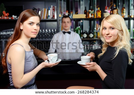 Coffee time. Two beautiful women drinking coffee and smiling to the camera while barman in white shirt and bow tie standing behind the counter