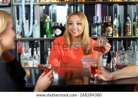 Talk at the bar. Smiling attractive female bartender talking to the guests holding a bottle with red drink