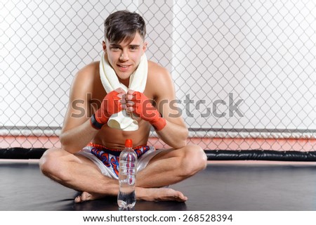 Short brake. Young smiling sportsman sitting with his towel on shoulders and a bottle of water in a fighting cage