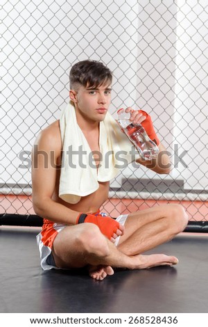 Water balance. Young kickboxer having a sip of water with a towel on his shoulders sitting in a fighting cage