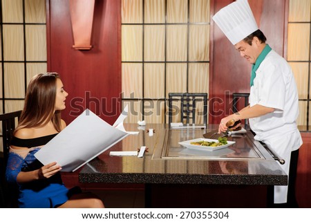 Professional consultation. Young attractive woman wearing a dress and holding menu copyspace is asking a cook while he cuts vegetables on a plate