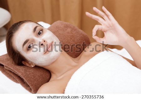 Perfect beauty service. Young woman showing ok to the camera when relaxing in a beauty parlor with a white mask on her face