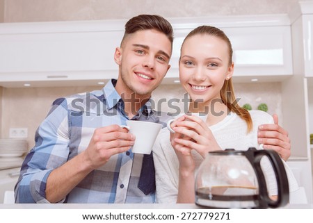Happy together. Two young smiling persons sitting next to each other in kitchen and drinking coffee