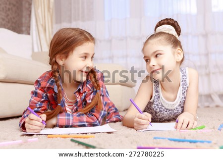 Eye contact. Cute smiling girls lying on carpet and drawing with help of colorful crayons and looking to each other