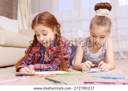 Devoted to work. Two serious little girls lying on carpet and drawing with help of colorful crayons and markers