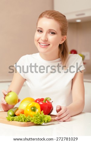 Look here. Smiling lady sitting at the table with cucumber pepper carrot and lettuce on her slicing board