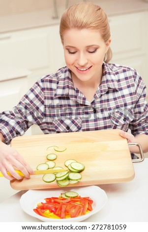 Being in high spirits. Young woman making salad of fresh vegetables and mixing sliced cucumbers with red and yellow pepper