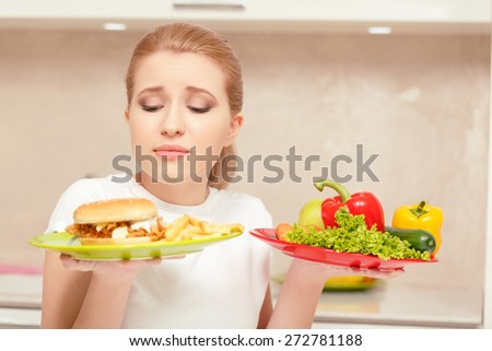 Feeling embarrassed. Beautiful lady holding plate with fast-food in one hand and vegetables in another feeling temptation to eat sandwich