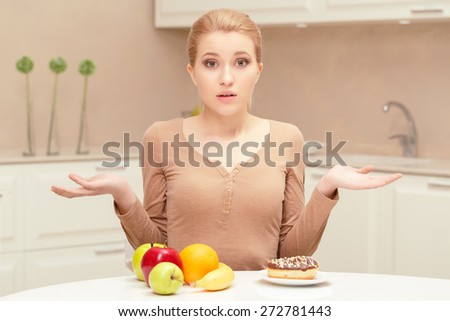 Difficult choice. Young pretty woman confused and hesitating sitting between fruit and plate with donut on her table trying to choose right food