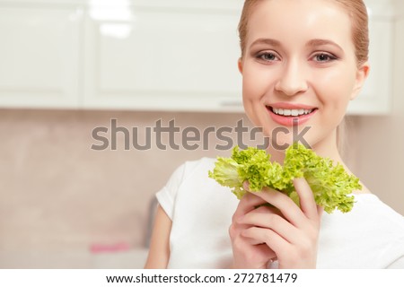 Feeling happy. Broadly smiling young woman standing in front of her kitchen cupboard, holding  bright green salad in her hands