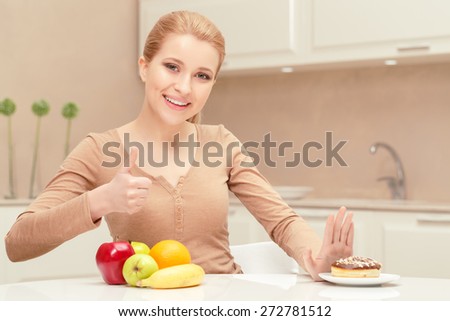 Made the right choice. Young lady sits at the table showing thumb up because she has strong will to decline eating donut and prefers apples and oranges