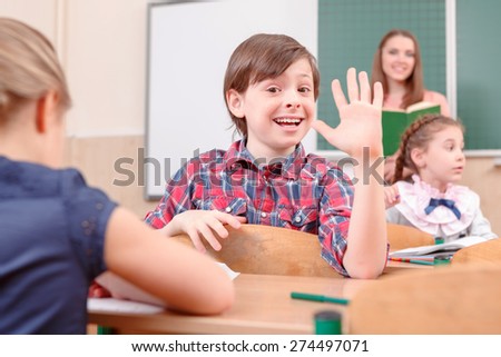 Hey everyone. Happy pupil boy waving with hand turned away from teacher in classroom.