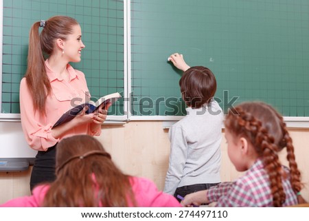 Ready to write. Young female smiling teacher and pupil standing at blackboard, pupil is going to write on blackboard.