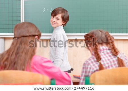 Smart child. Little smiling boy standing near green blackboard in classroom.
