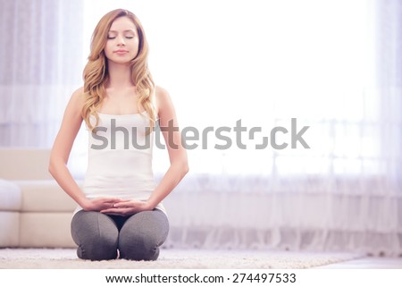 Stay positive. Young beautiful lady sitting on white carpet on her knees with closed eyes, crossed hands and meditating.
