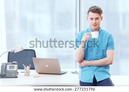 In high spirits. Young smile businessman sitting on desk covered with office equipment with cup of coffee.