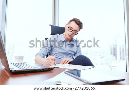 Attentive writing. Young handsome businessman sitting at desk covered with notebook papers and tablet filling papers in office.