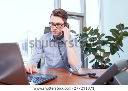 Concentrated attention. Young businessman sitting at desk working on computer and making calls in office.