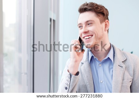 Pleasant conversation. Young smiling handsome man talking per mobile phone in front of window.