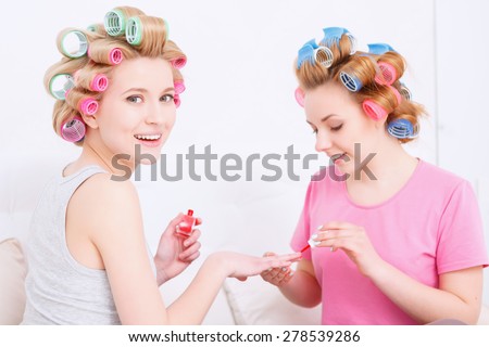 Portrait of young girls wearing pajamas and colorful hair rollers painting nails in red color and smiling at pajama party in the light room