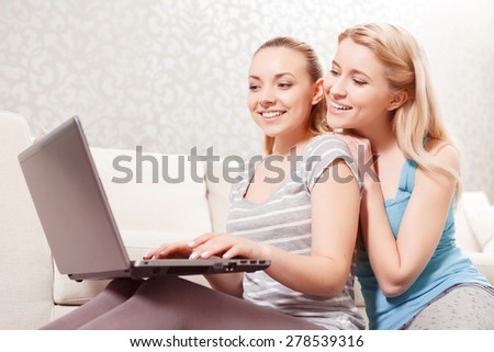 Friendship. Two young beautiful blond girls wearing pajamas sitting on the couch and looking at laptop